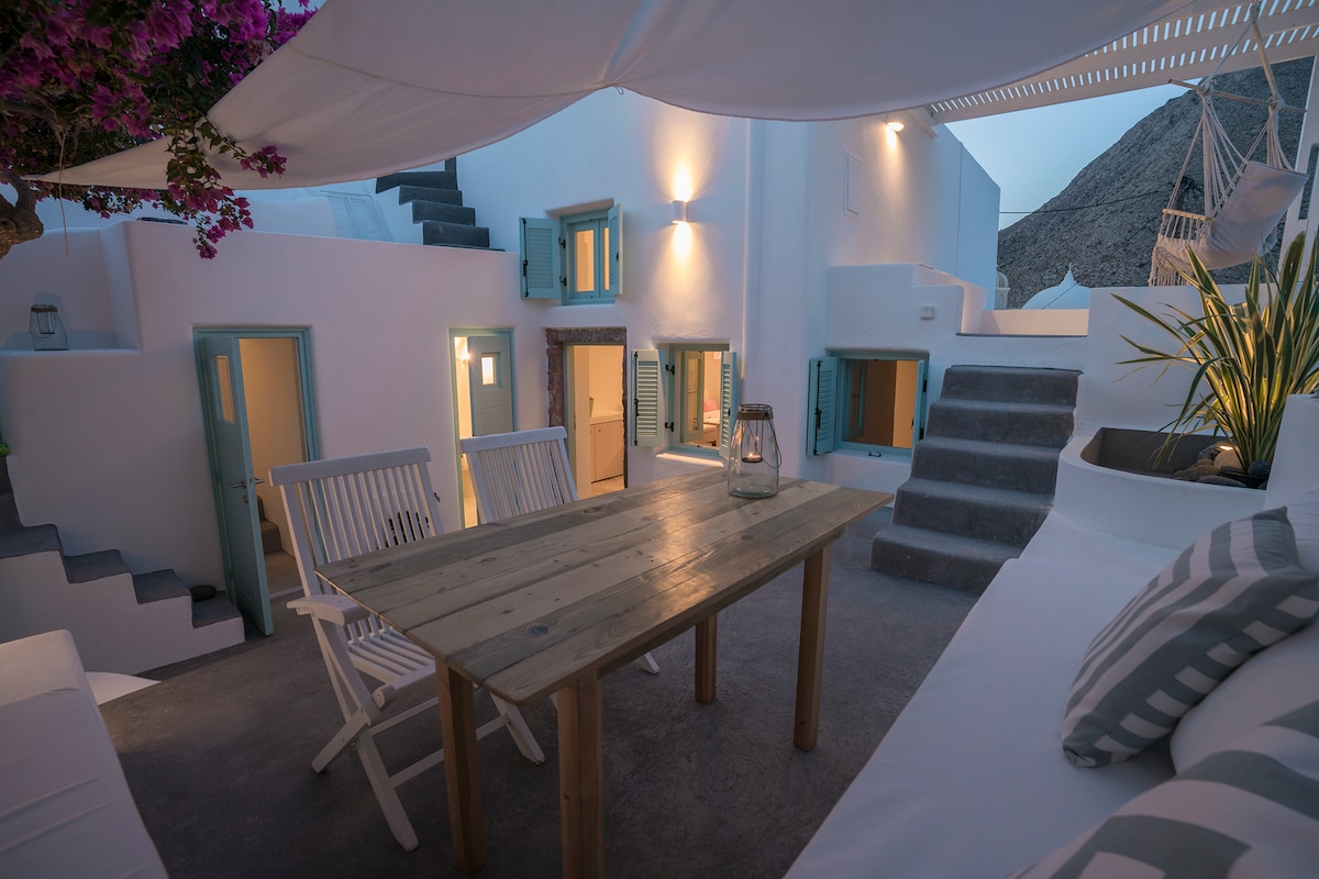 An outdoor patio area is captured at dusk, featuring a wooden dining table surrounded by white chairs. Soft lighting illuminates the space, enhancing the white-washed walls and the surrounding greenery. A stairway leads to various doorways, inviting exploration of the cave-style rooms.