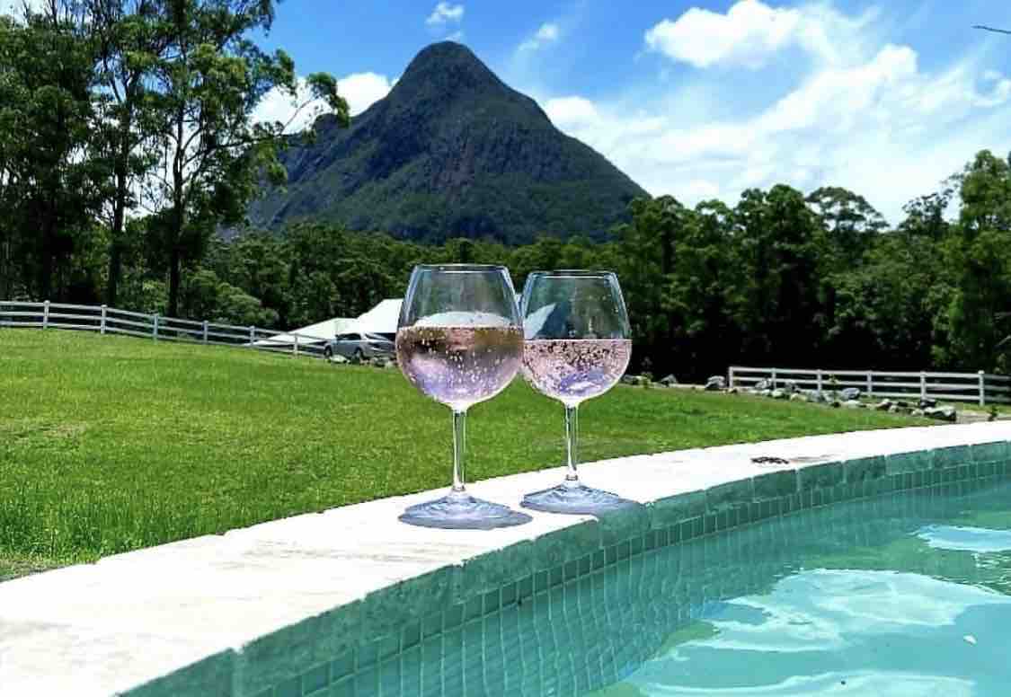 Two glasses filled with sparkling beverage sit on the edge of a pool. The background features a large mountain amidst lush greenery, with a white fence framing the expansive lawn that leads to additional buildings in the distance.