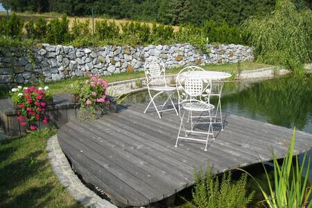 A wooden deck extends over a tranquil swimming biotope, featuring a round table with two white chairs. Lush greenery and colorful flowers are present, complementing the serene setting framed by a low stone wall in the background.