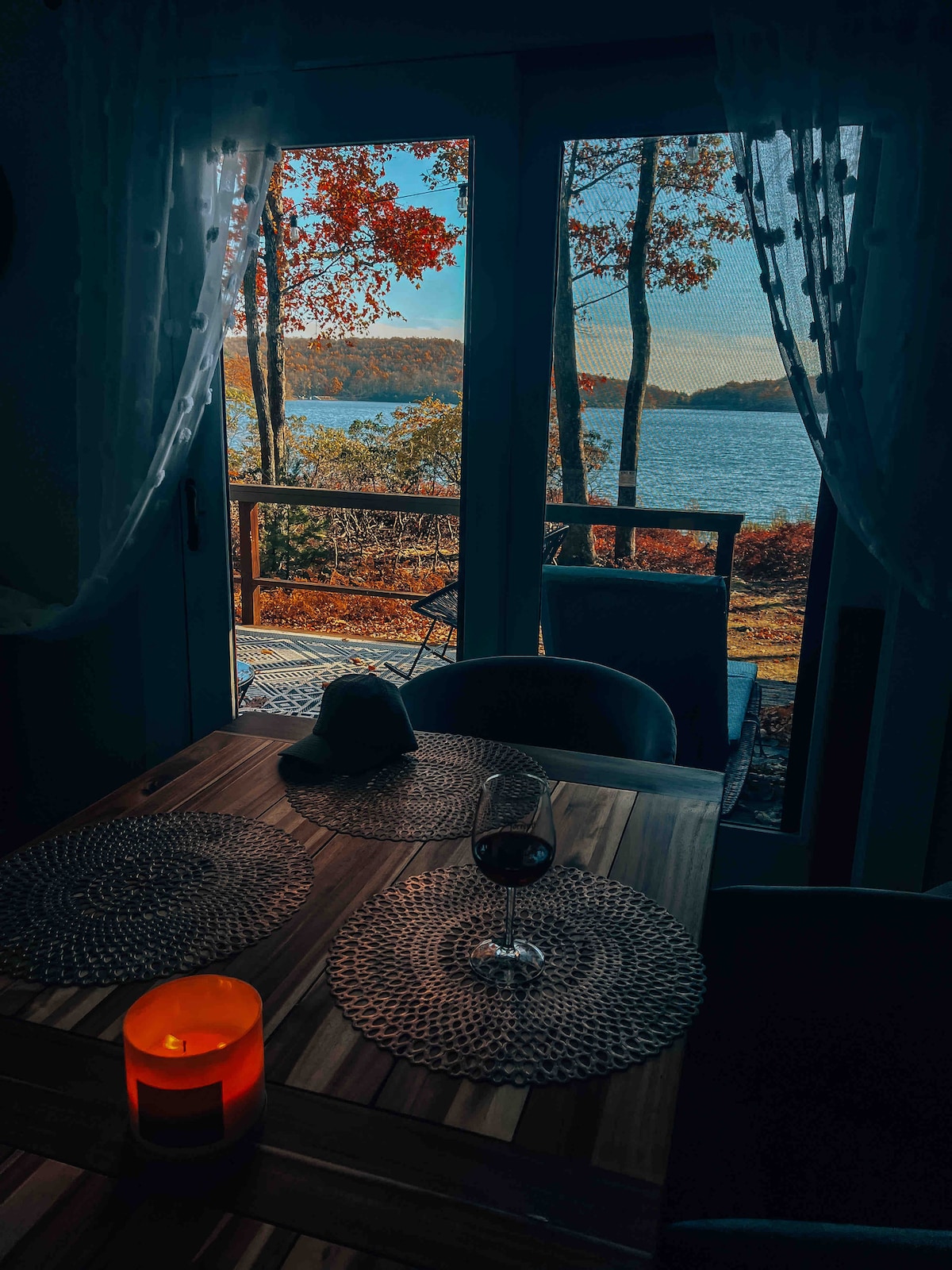 A dining area is visible, featuring a polished wooden table set with woven placemats. A candle glows softly next to a glass of red wine. Large glass doors open to a scenic view of fall foliage and a serene lake.