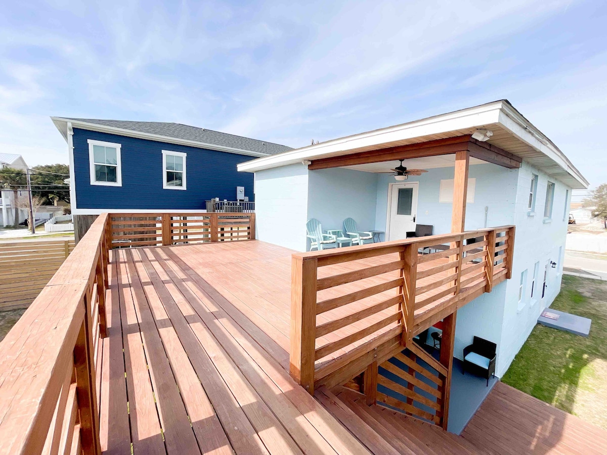 A large private deck made of polished wooden planks spans the upper floor unit, providing ample space for seating. The structure features a light blue exterior, complemented by a blue house next door. A ceiling fan is visible, enhancing airflow in the outdoor area.