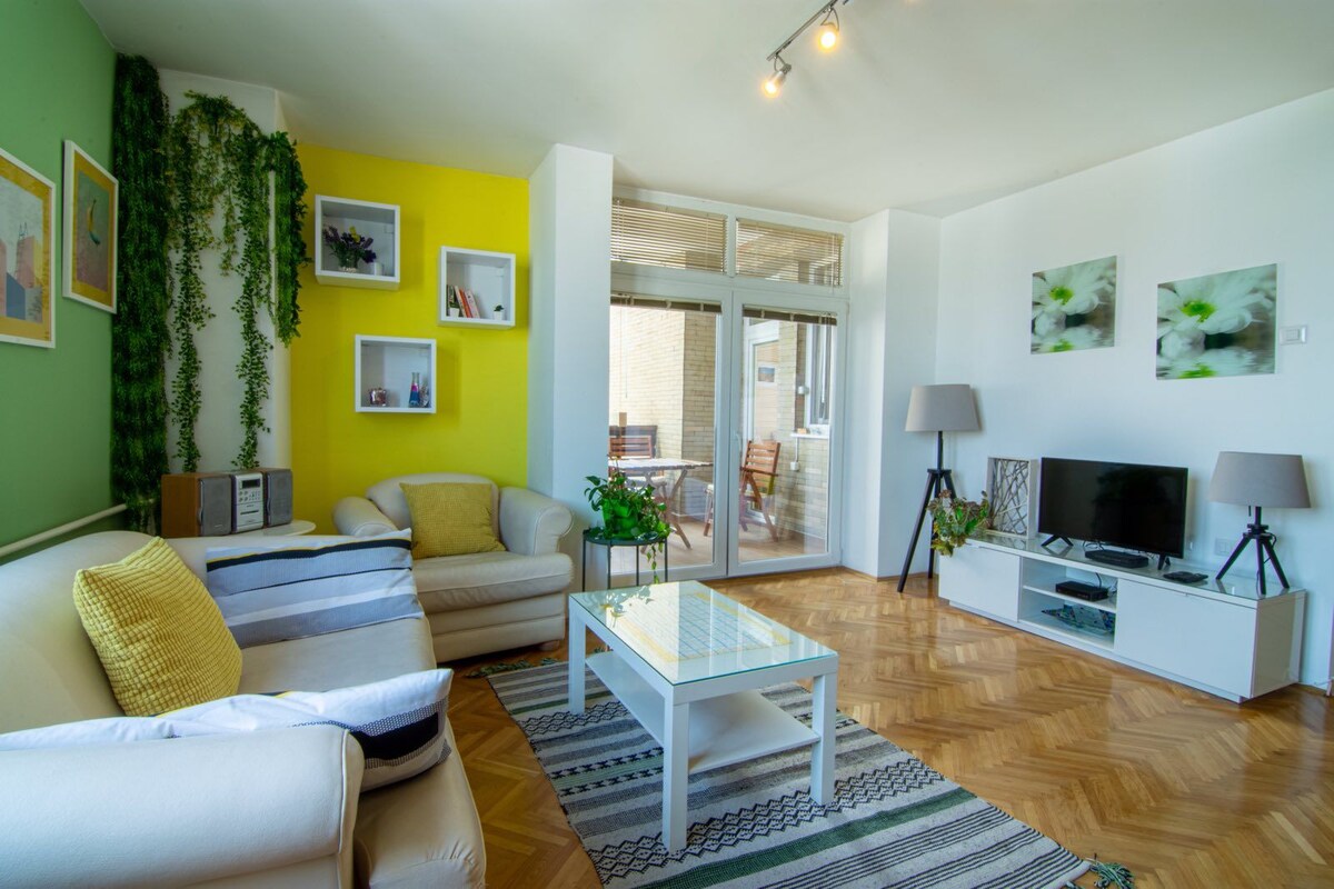 The living area features bright green and yellow walls, complemented by a white sofa and armchair. A coffee table sits on a patterned rug. Shelves display various decorative items. A television is positioned above a low-lying cabinet, and a large window offers access to the terrace.