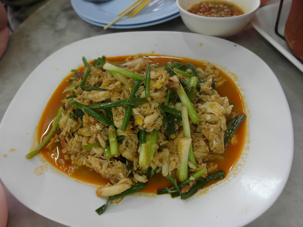 A plate of vibrant stir-fried vegetables is presented, featuring chopped green onions and tender pieces of chicken. The dish is coated in a rich, savory sauce, with steam rising gently from the surface, indicating warmth. A small bowl of dipping sauce is visible in the background.
