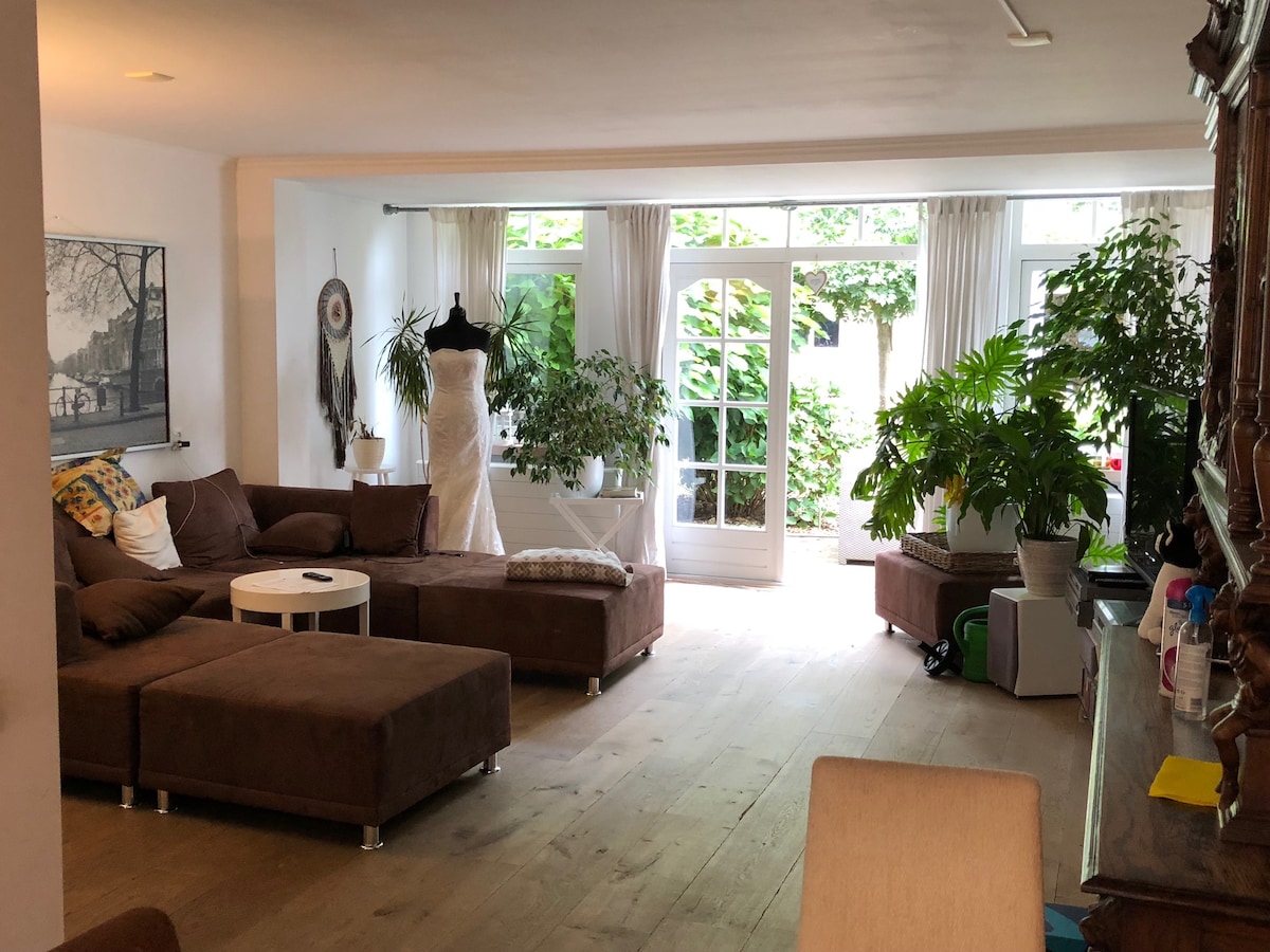 A spacious living room is depicted, featuring a large brown sectional sofa and a coffee table. Natural light floods the area through glass doors that open to a garden, surrounded by potted plants. The room includes a wall art piece and is designed for comfort and relaxation.