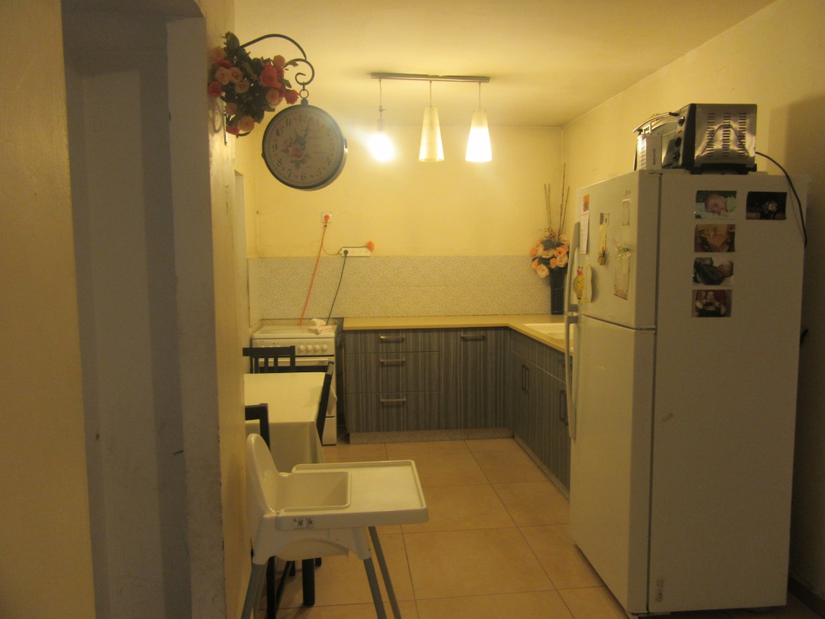 A compact kitchen area features a light-colored countertop and modern cabinetry with a gray finish. Overhead pendant lights illuminate the space. A high chair is positioned beside a small dining table, while a refrigerator showcases various magnets on its surface.