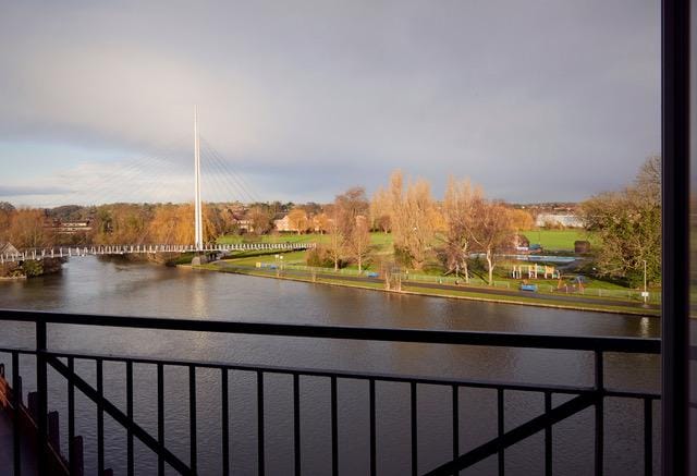 A scenic riverside view is visible from the apartment's Juliet balcony, showcasing the river reflecting the sky. Lush greenery and trees line the riverbank, with a park area featuring walking paths and playgrounds. A bridge spans the river in the distance, enhancing the tranquil setting.