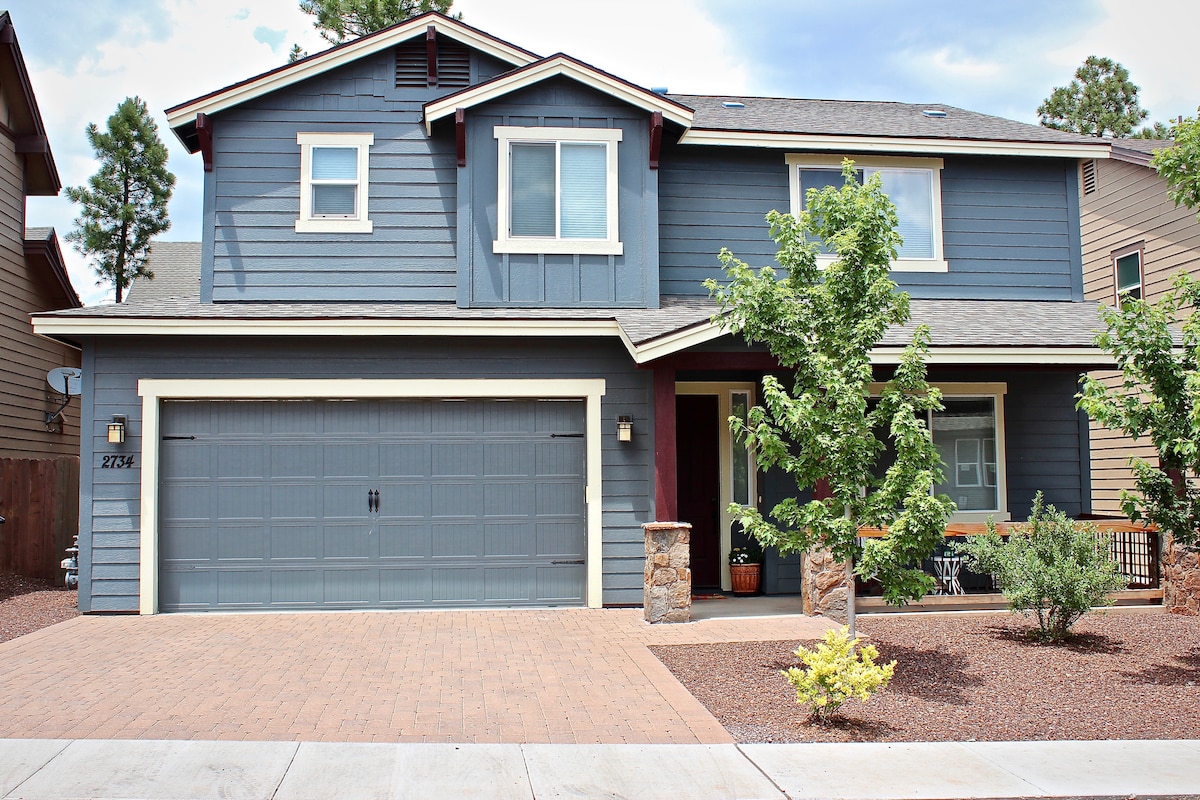 The exterior of a two-story home features a blue siding with white trim and a welcoming front porch. A stone wall accents the garage, which has double doors. Landscaped areas include small shrubs and trees, set against a backdrop of a clear sky.