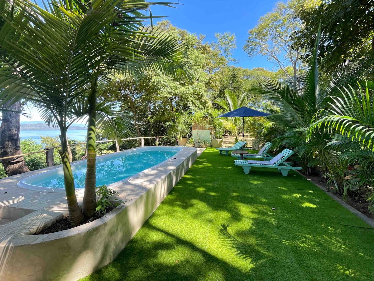 A serene outdoor space features a refreshing pool surrounded by lush greenery. Comfortable lounge chairs are positioned nearby under a blue umbrella, and sunlight creates a warm ambiance throughout the area. The view extends towards the ocean, enhancing the peaceful setting.