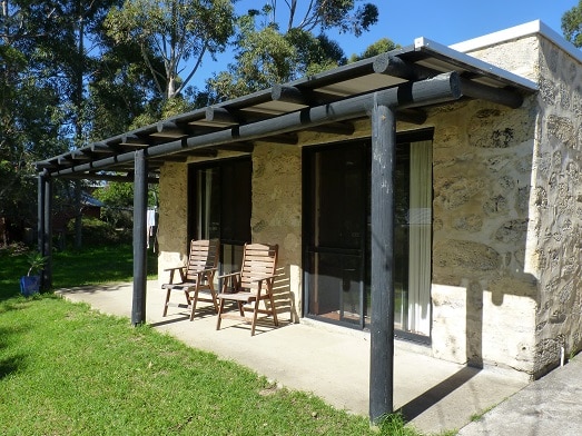 The self-contained limestone cottage features a shaded porch with two wooden chairs placed against a natural stone facade. Lush green grass surrounds the entrance, complemented by tall eucalyptus trees in the background, creating a serene outdoor setting.