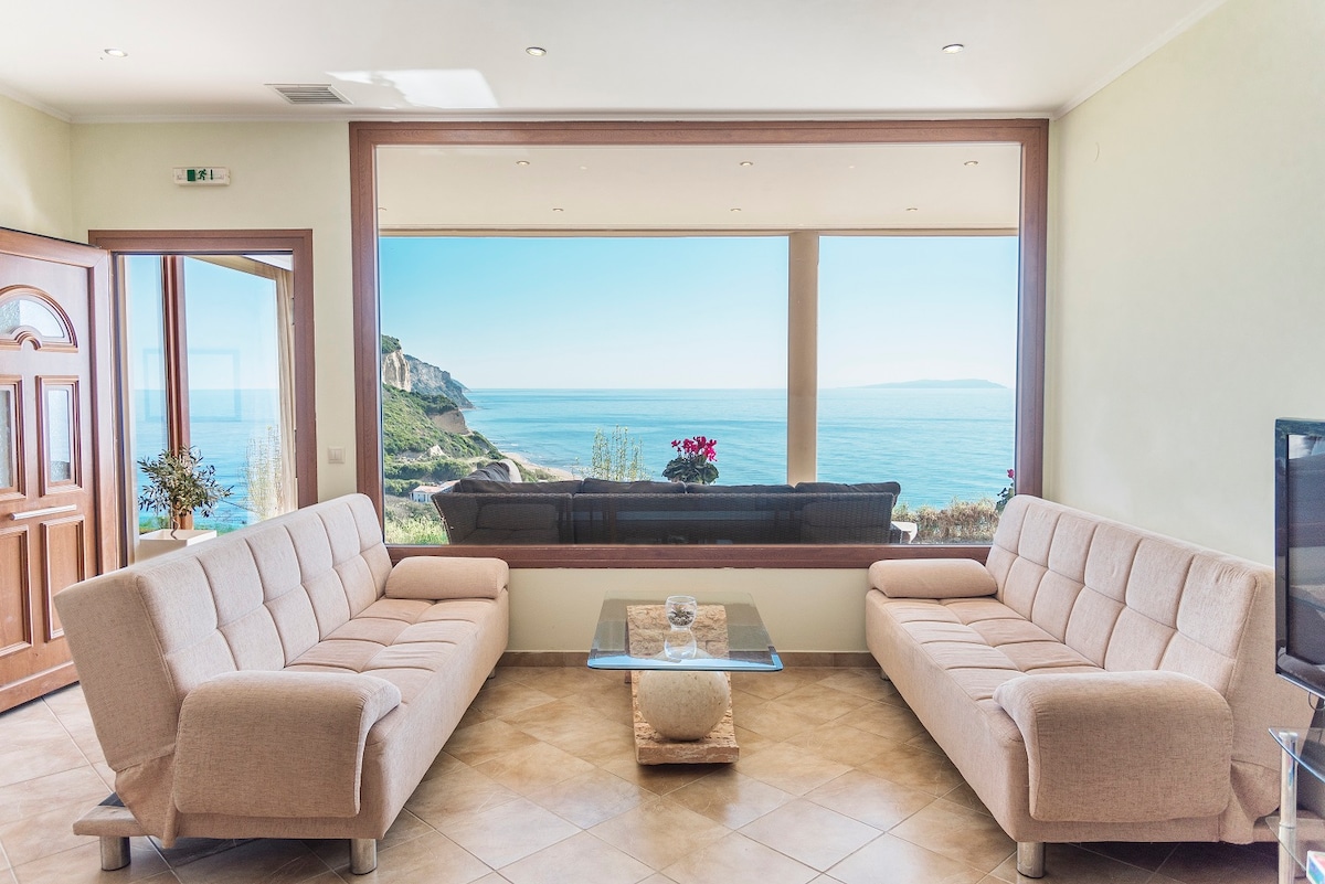 A bright living area features two comfortable sofas positioned around a glass coffee table. A large window frames a panoramic view of the sea, with scenic hills visible in the background. Natural light enhances the airy atmosphere of the space.