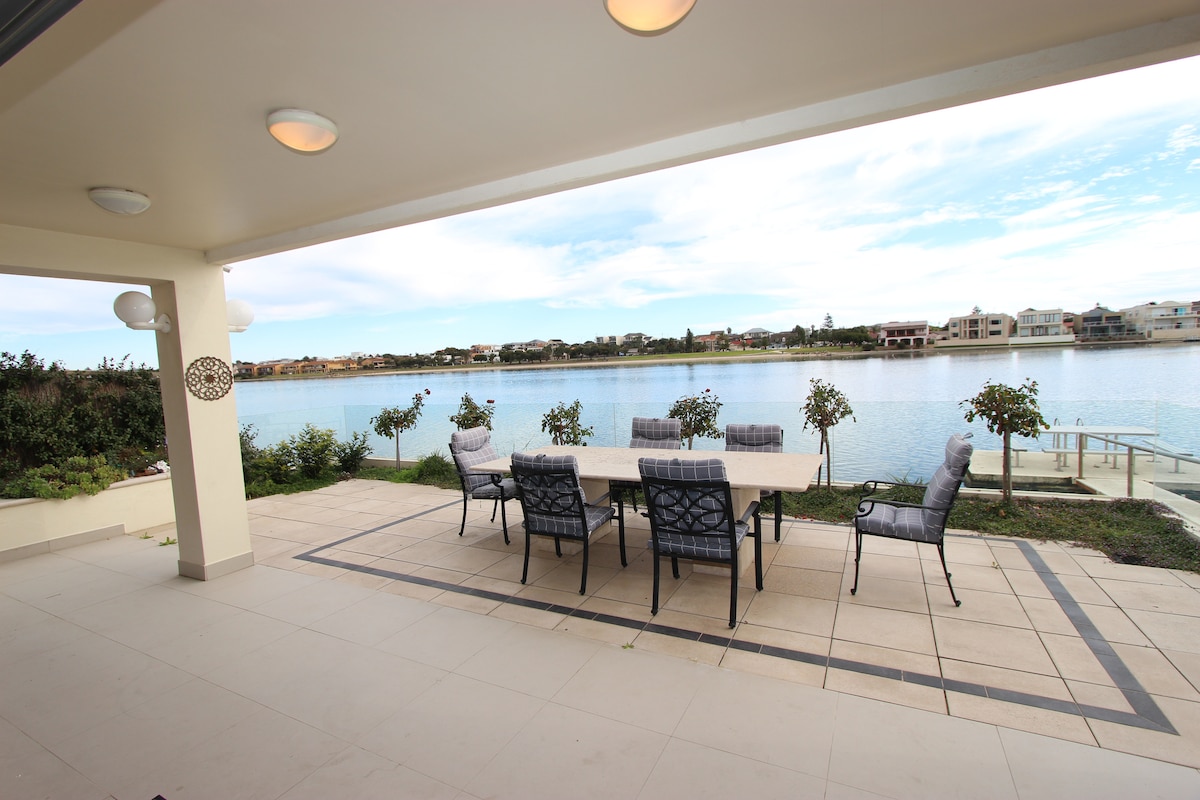 A spacious outdoor dining area is positioned on a tiled patio, overlooking a calm lake. The table is surrounded by several cushioned chairs, with greenery and water visible in the background. Soft lighting fixtures complement the serene atmosphere.