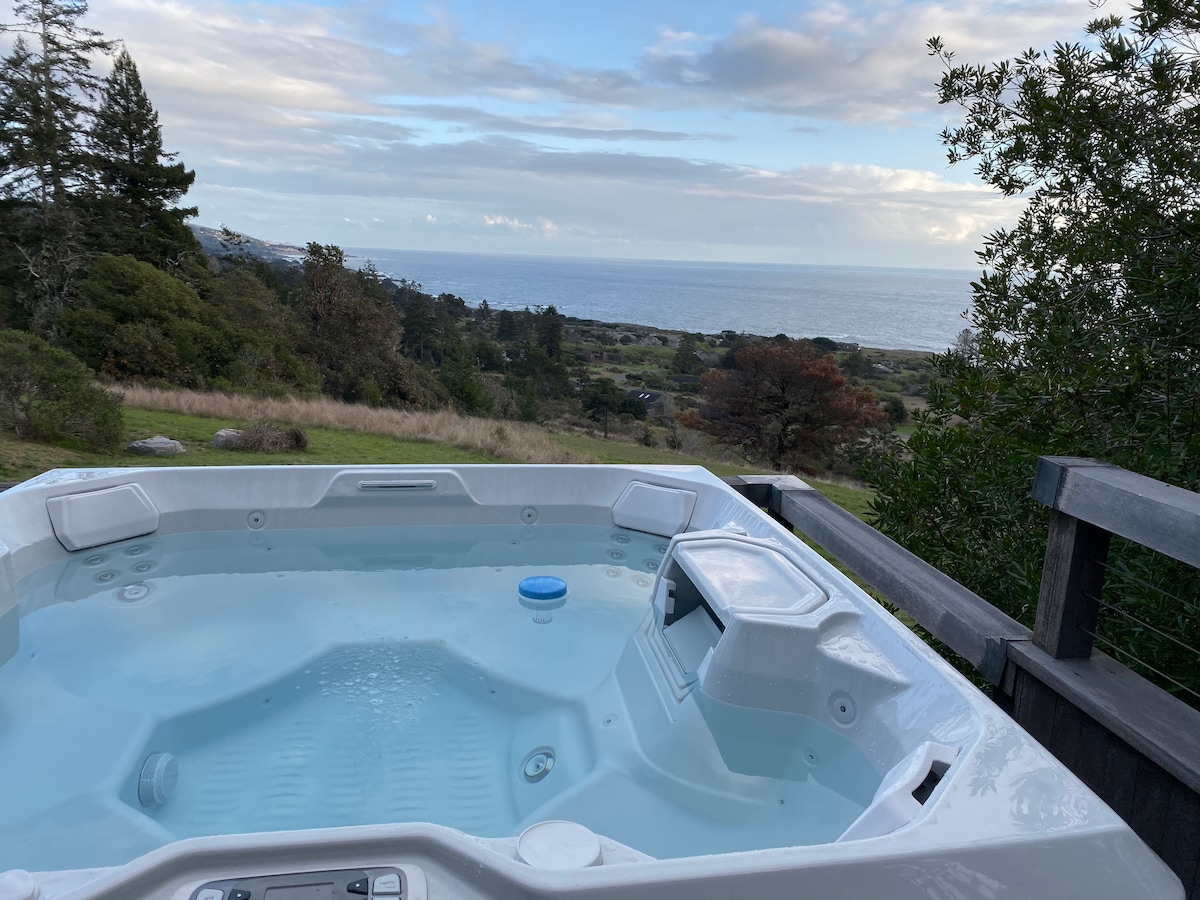 A private hot tub is positioned on a deck, offering views of the Pacific Ocean and the surrounding landscape. The water's surface reflects the sky, while the lush greenery frames the scene. The calming atmosphere is enhanced by the vast, unobstructed horizon.
