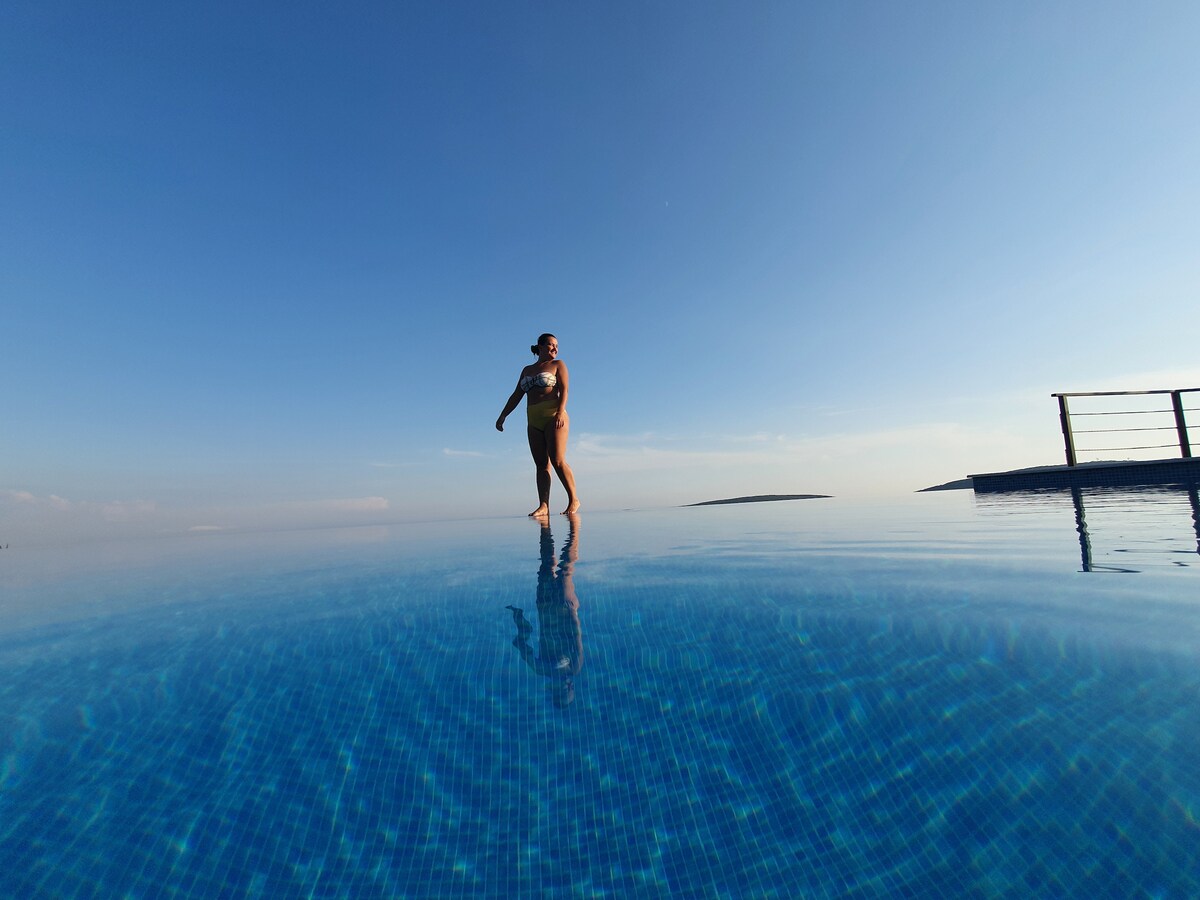 An infinity pool extends towards the horizon, reflecting the clear blue sky. A person is seen walking along the edge of the pool, while distant land is visible in the background, contributing to a serene atmosphere.
