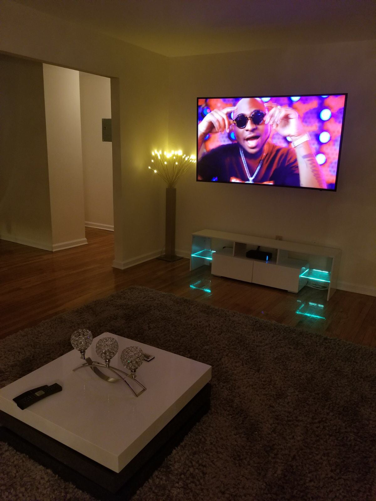 A cozy living room features a modern television mounted on the wall, illuminated by a soft glow from a decorative floor lamp. A stylish coffee table with a minimalist design is centered on a plush area rug, creating a welcoming gathering space.