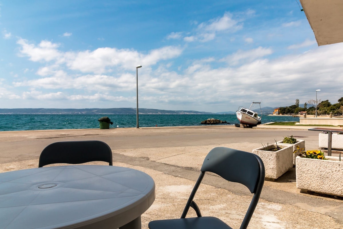 A view of the sea is captured from a waterfront area, featuring a circular table with two blue chairs positioned nearby. A small boat rests on the shore, and distant hills are observable under a partly cloudy sky. The shoreline is lined with simple planters.