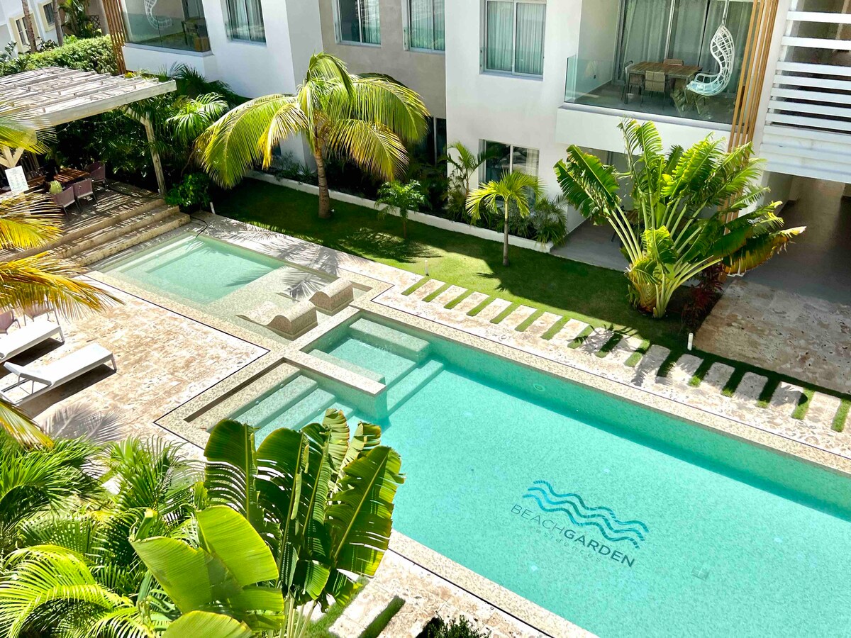A serene communal pool area is surrounded by lush greenery and tropical plants. The pool features distinct sections with clear turquoise water, complemented by a stylish lounge area with comfortable seating positioned nearby. The structure's modern design is reflected in the surrounding greenery.