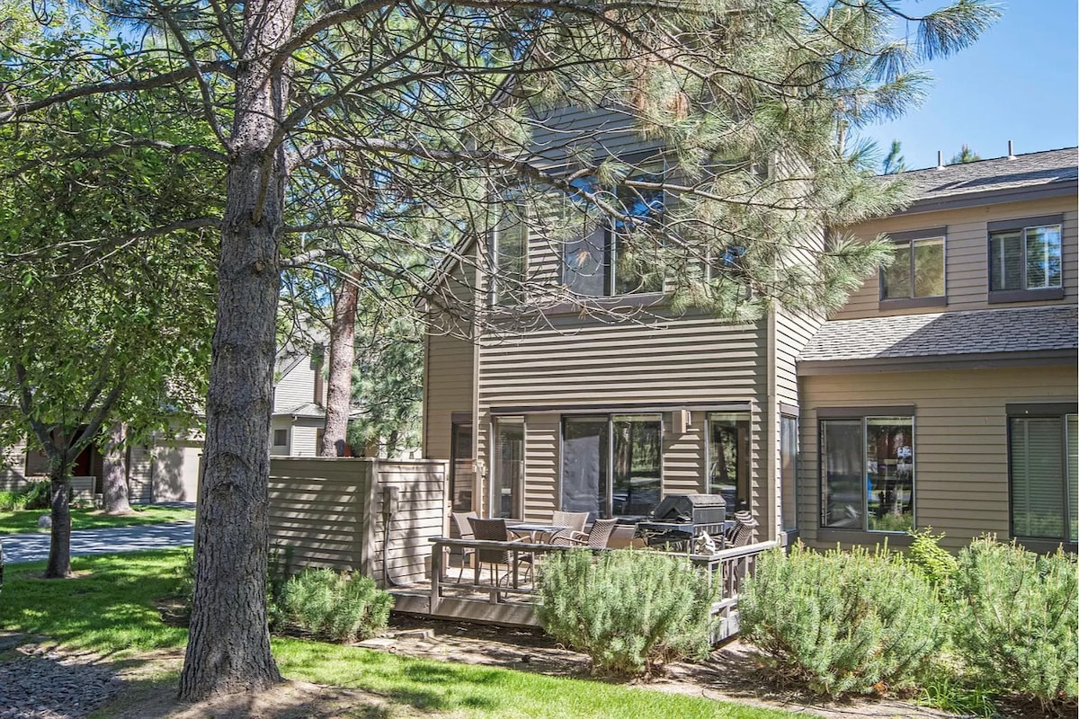 A two-story townhouse is surrounded by trees, featuring a private deck adorned with outdoor seating. Large windows illuminate the exterior, showcasing a contemporary design that blends with the natural landscape.