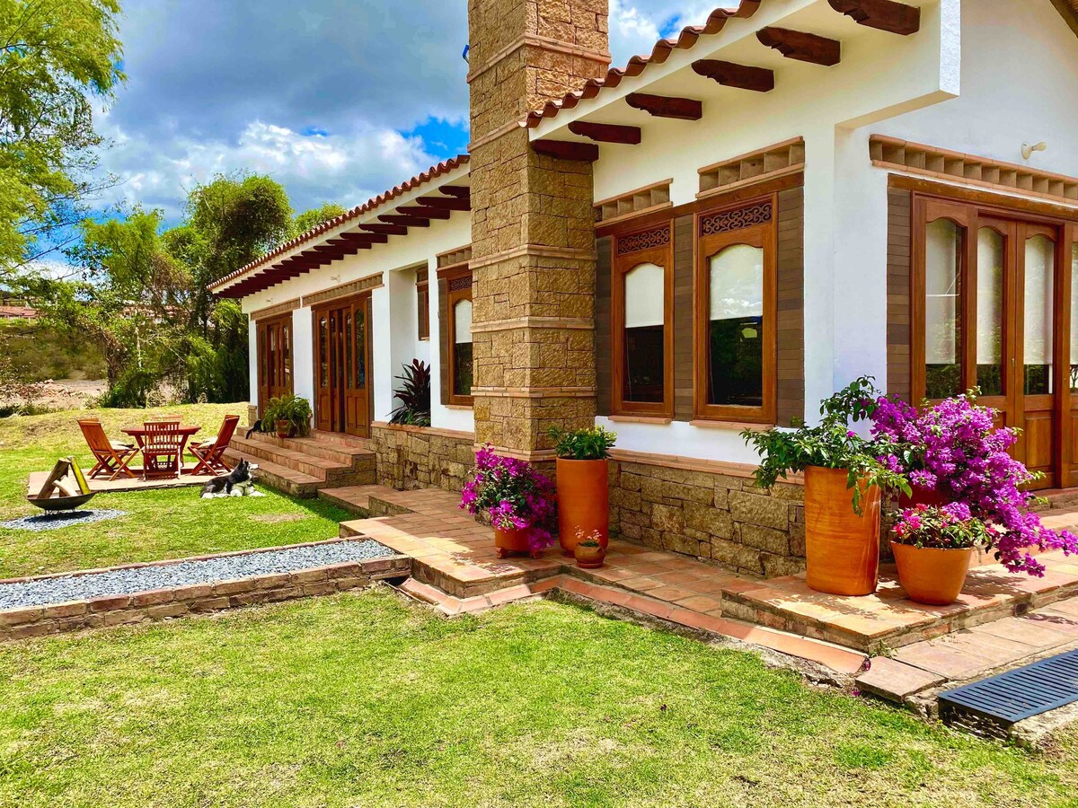 A charming outdoor space features a well-maintained lawn and terracotta planters filled with vibrant flowers. A rustic stone wall supports the structure, while wooden doors and windows enhance the facade. A dining area with chairs and a table is positioned on a stone patio.