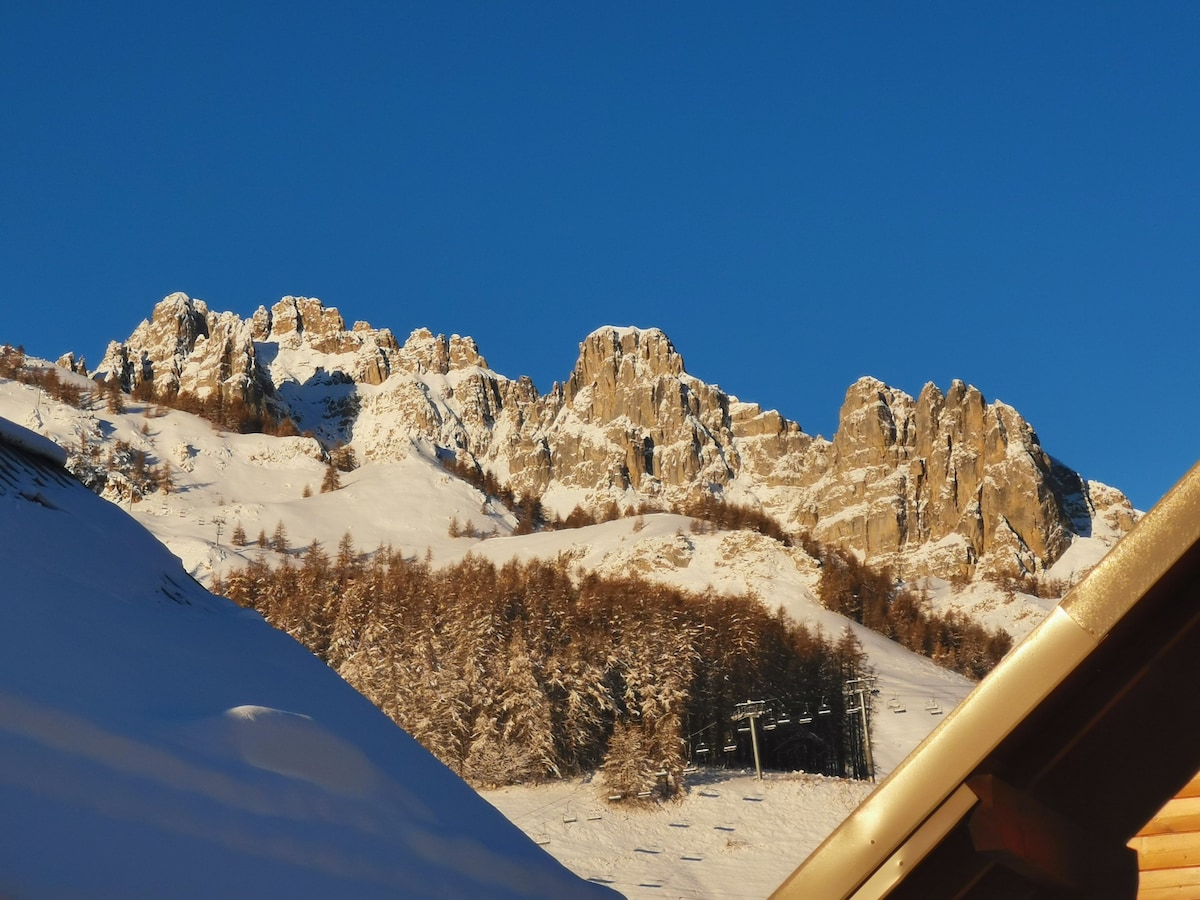 Snow-covered mountains under a clear blue sky are visible, showcasing rugged peaks and a pine tree line below. The image captures the winter landscape of the Réallon ski area, with ski lifts subtly located in the foreground.