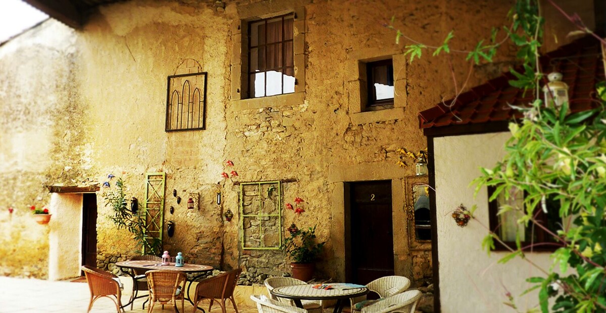 A rustic courtyard area features a textured stone wall, adorned with potted plants and framed art. A round table with wicker chairs is situated under a shaded area, inviting relaxation. Windows with wooden frames overlook the space, adding character to the charming outdoor setting.