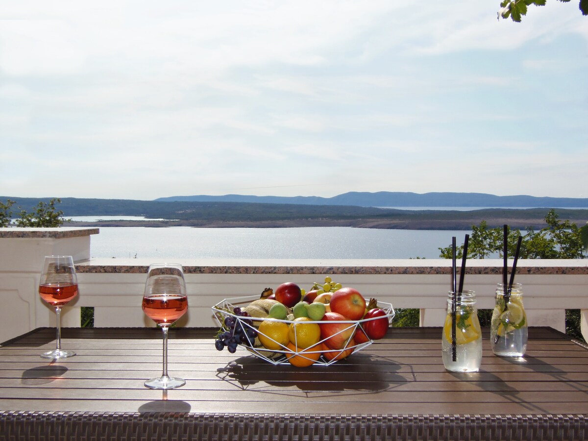 A dining table is set on a terrace, featuring a metal fruit basket filled with various fruits. Two glasses of rose wine and two glasses with straws and lemon slices sit on the table, with a scenic view of the sea and distant hills visible in the background.