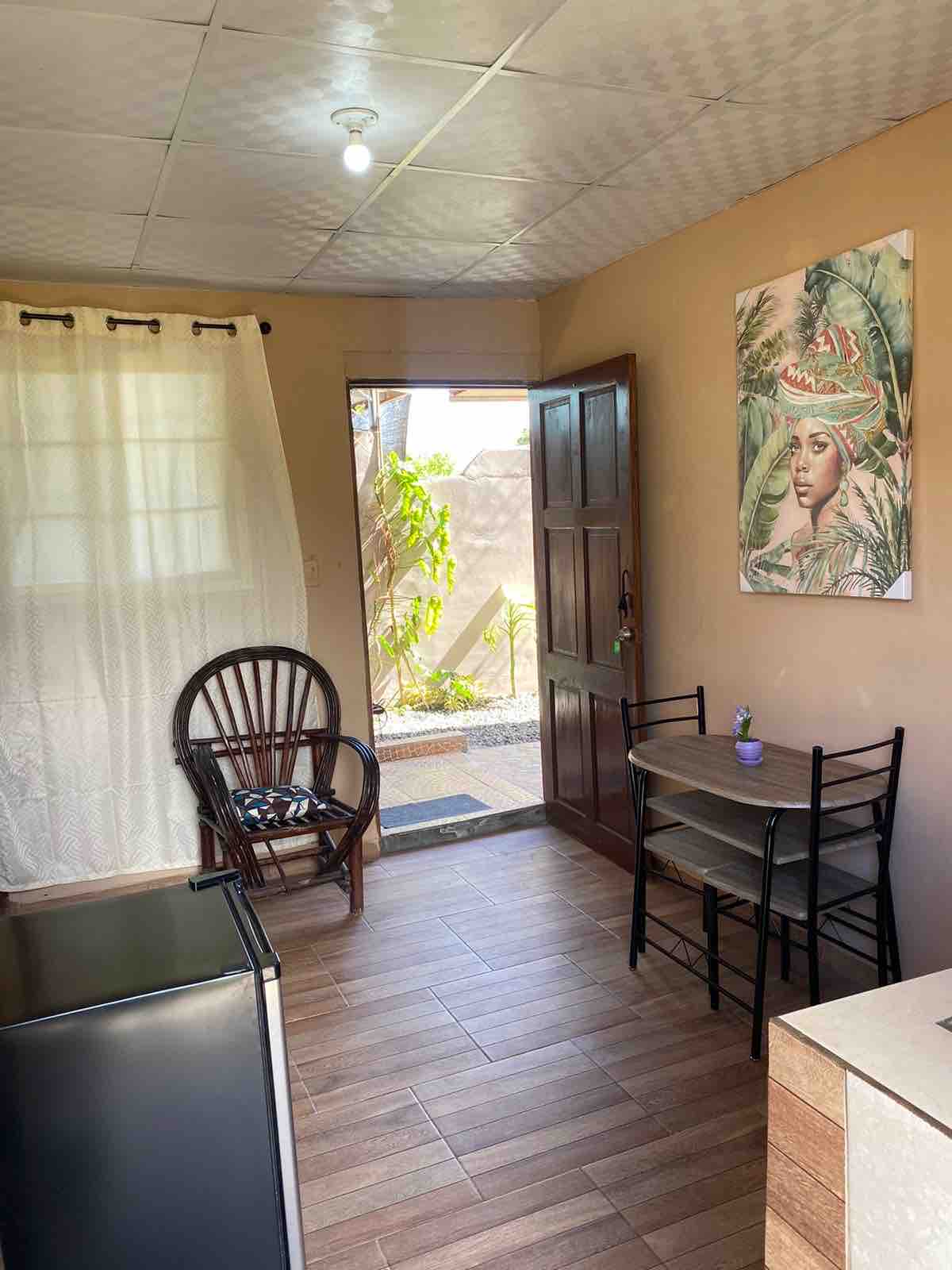 A small kitchen area is shown, featuring a round table with two chairs and a natural light source from the open door. A comfortable chair is positioned near the window, and a refrigerator is visible on the left. Decor with leaf prints is seen on the wall.