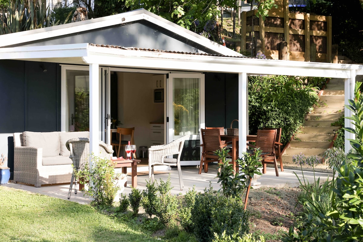 A shaded outdoor area features comfortable seating options, including plush sofas and wooden dining furniture. Lush greenery surrounds the space, providing a sense of privacy. A pathway leads up to the property, with steps visible in the background.