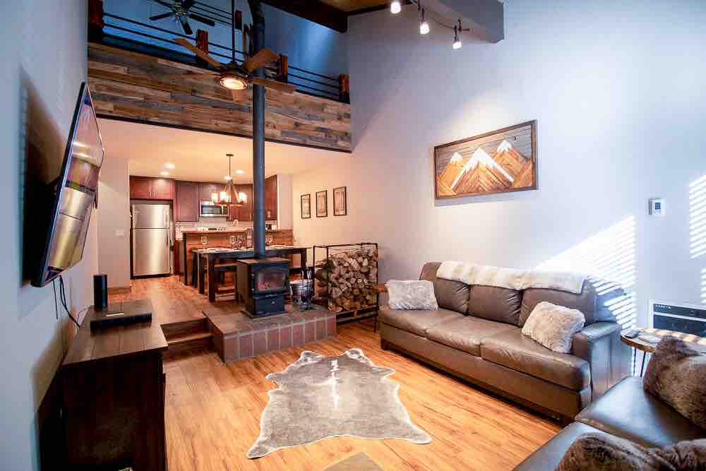 The open-concept living area features a cozy seating arrangement with a leather sofa and a loveseat, alongside a functional fireplace. A 65" TV is mounted on the wall. Natural light filters in through high windows, accentuating rustic wood details and a mountain-themed artwork.