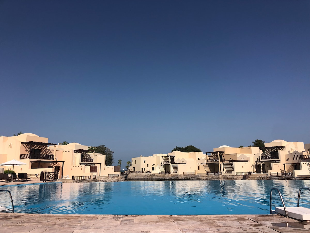 A clear blue sky complements the expansive adult pool, reflecting the surrounding architectural structures. The resort's buildings, designed with soft, neutral tones, are visible in the background, enhancing the serene atmosphere of the area.