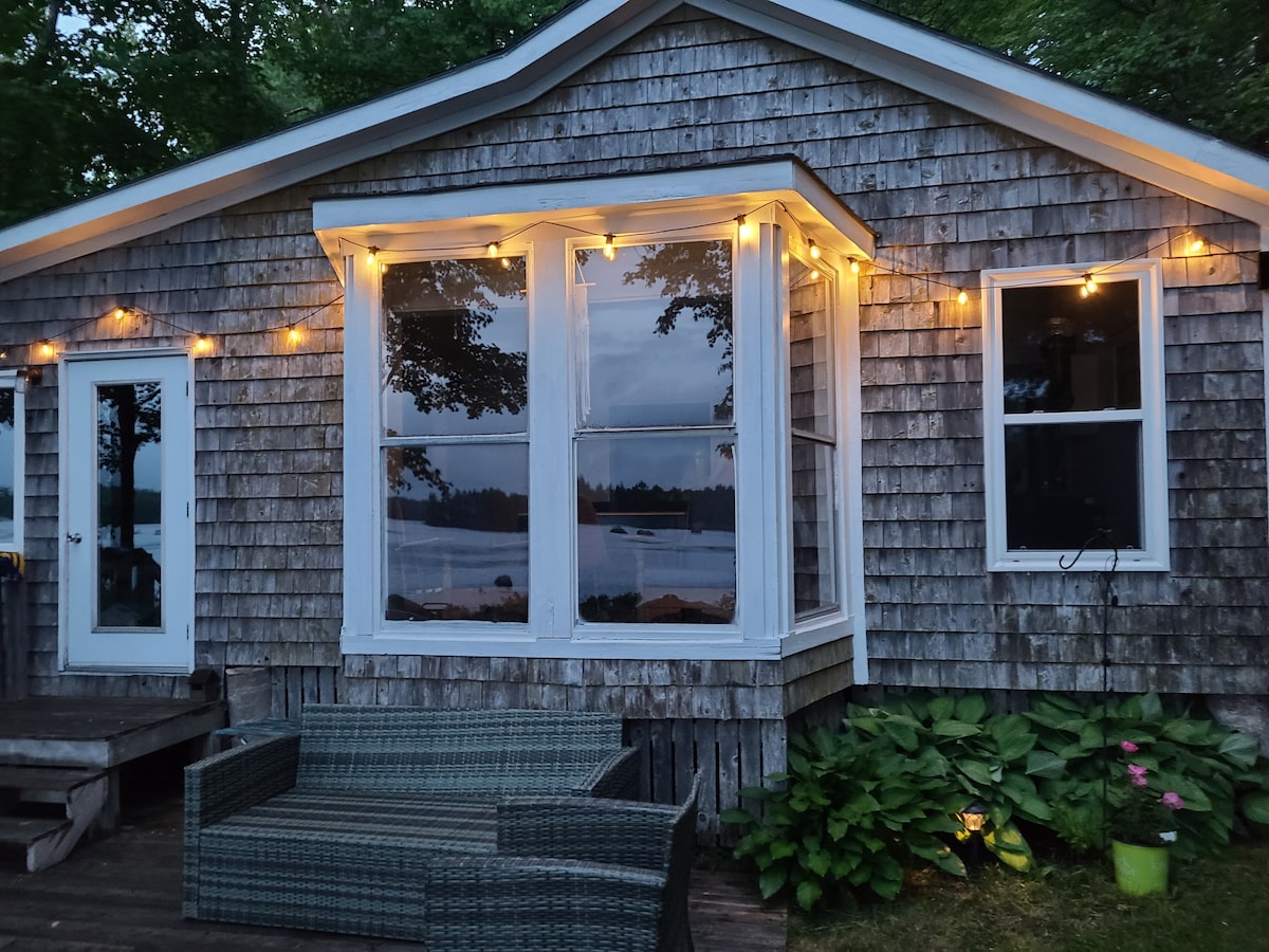 The exterior of a cottage displays natural wood siding and large windows, providing views of the lake. String lights illuminate the area, enhancing the welcoming ambiance. A comfortable seating arrangement is visible on the deck, surrounded by lush greenery and a quiet lakeside setting.