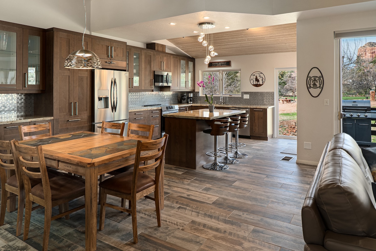 An open-concept kitchen and dining area features modern appliances and warm wooden cabinetry. A rustic dining table with seating for six is positioned beside a sleek kitchen island with bar stools. Natural light streams through windows, highlighting views of the outdoor space.