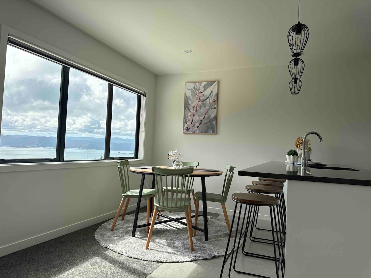A dining area features a round table surrounded by four green and black chairs, with a soft area rug underneath. Large windows provide a view of the landscape, while a decorative plant and artwork add a touch of elegance to the space.