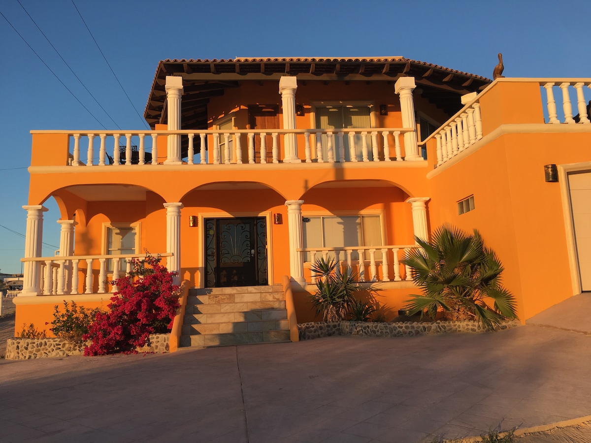 A vibrant orange two-story home is framed by white columns and a terracotta roof. Lush greenery and blooming pink flowers adorn the entrance, which is accessed by a set of stone steps leading to a decorative front door.