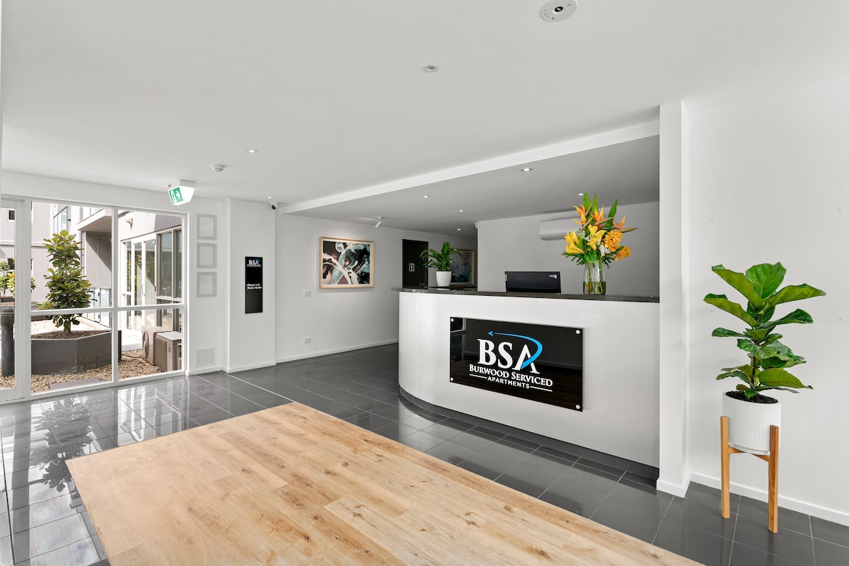 The reception area features a modern design with a large, sleek front desk displaying the property name. Bright yellow flowers are arranged on the counter, while indoor plants add greenery. Large windows provide natural light, illuminating the polished floor and minimalist decor.