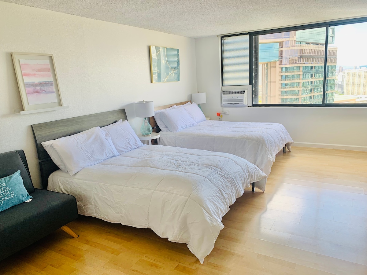 A well-lit studio space is presented with two spacious beds covered in white linens. A modern gray headboard is featured along with two bedside lamps. A small seating area is visible, and large windows offer views of the outdoor skyline.