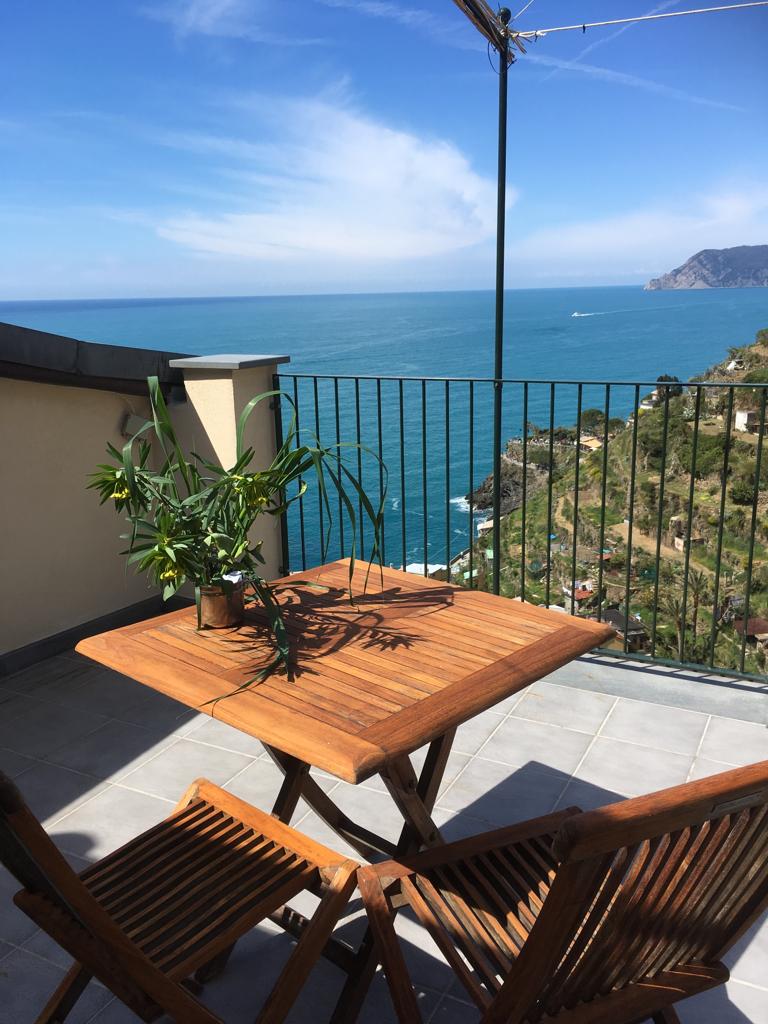 A wooden table and two chairs are positioned on a terrace that overlooks the sea. The expansive view showcases vibrant blue waters and rugged coastline, with hills in the background. A potted plant adds a touch of greenery to the table.