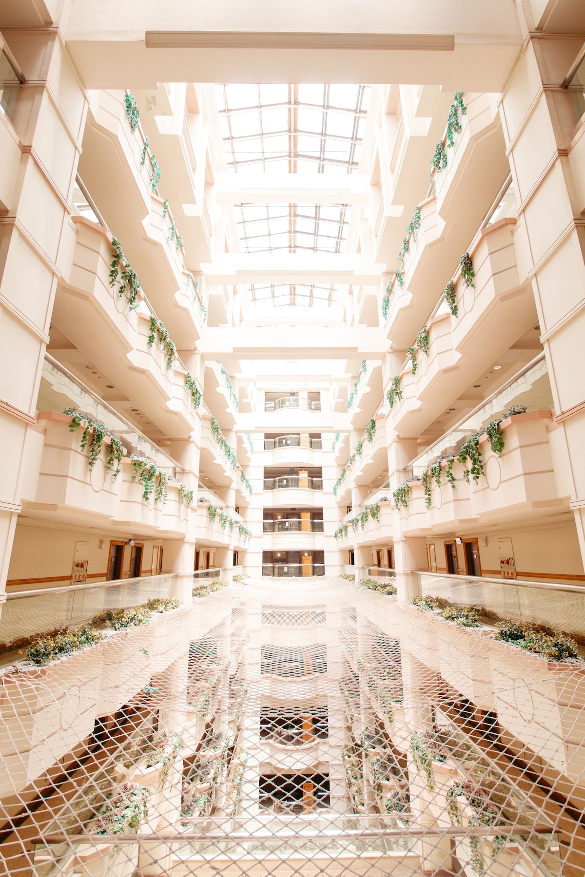 An airy atrium features multiple levels with open walkways lined by greenery. Natural light floods the space from a skylight above, illuminating the glossy surfaces below. A netted railing provides safety while allowing views into the central area, enhancing the open atmosphere.