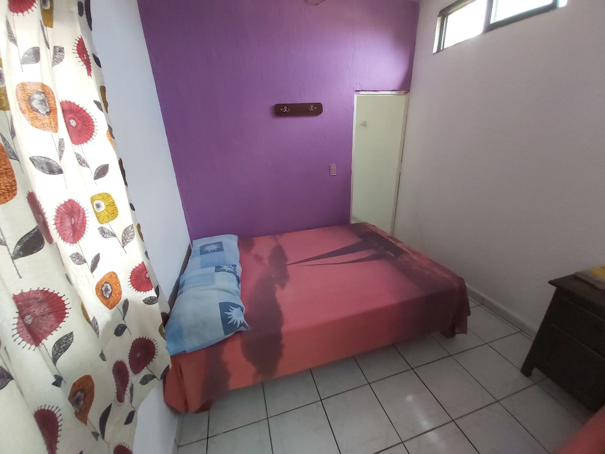 A sleeping area is displayed, featuring a spacious bed with a vibrant multicolored blanket. A light purple wall complements the bedding, while natural light enters through a pair of small windows. A rear doorway and a simple wooden nightstand are also visible.