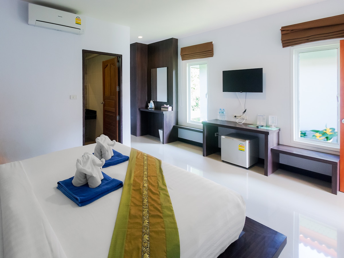 A spacious room features a large bed adorned with two neatly arranged towels. Sunlight streams through large windows, illuminating the bright interior. A flat-screen TV is mounted on the wall, and a desk with a mirror provides additional functionality. Air conditioning ensures a comfortable atmosphere.