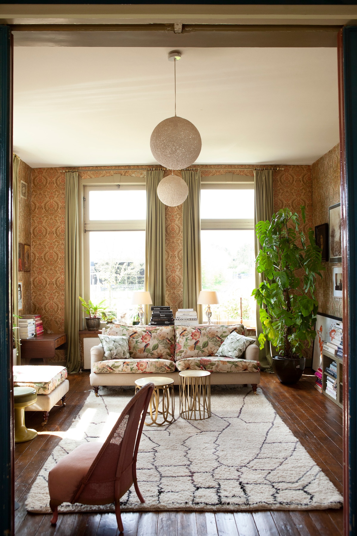 The living area features a floral-patterned sofa centered between two small tables, illuminated by natural light from large windows. A decorative plant adds greenery, and warm textiles are present on a light-colored rug, enhancing the cozy atmosphere.