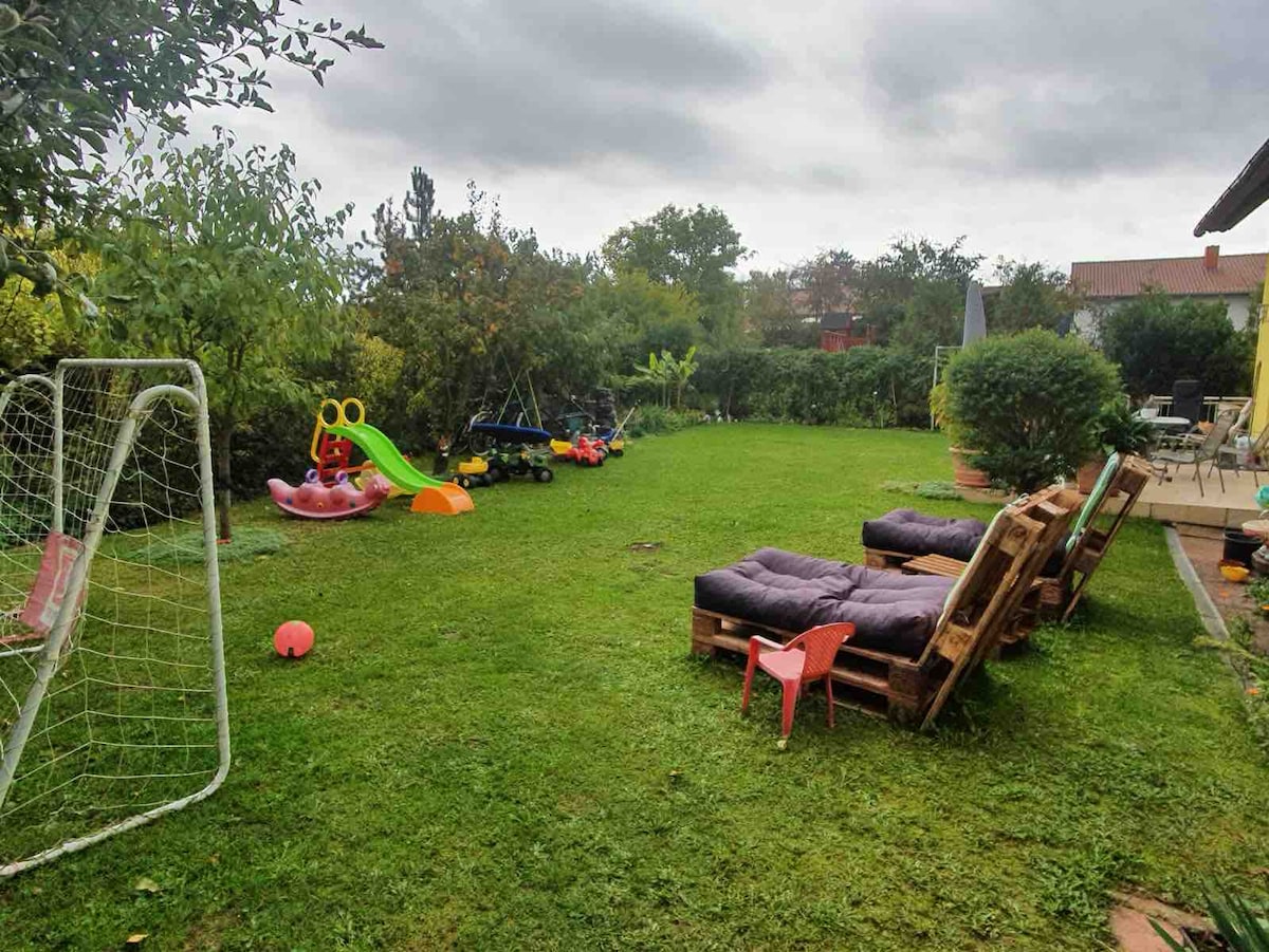 A spacious garden area is visible, featuring well-maintained grass and a variety of play equipment. Two lounge chairs made from wooden pallets are positioned on one side. A children's slide and ride-on toys are also present, accompanied by a soccer goal and colorful play items.