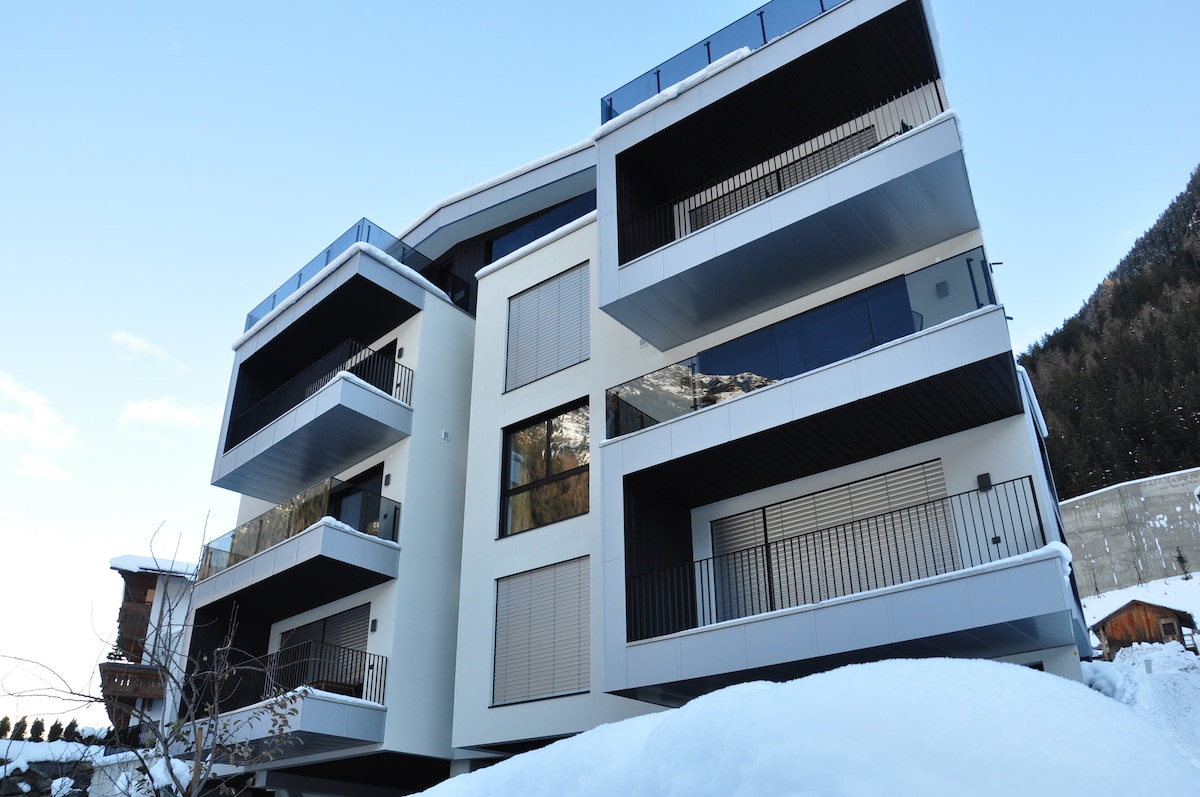 The modern building features multiple balconies, each showcasing sleek lines and expansive glass windows. The structure is set against a backdrop of snow, integrating harmoniously with the winter landscape. The upper levels' balconies offer views of the surrounding mountains.