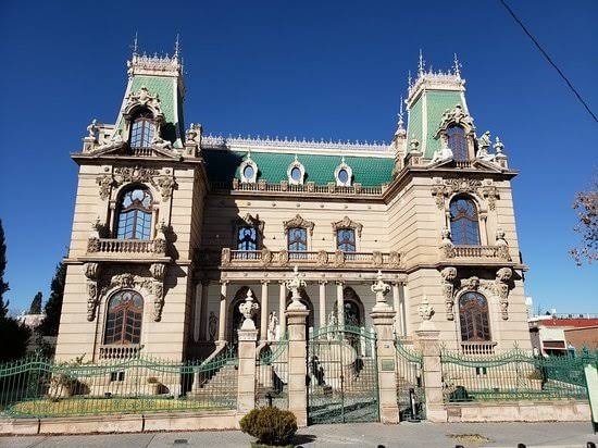 A grand, historic building features intricate architectural details and a distinctive green roof. The façade is adorned with ornate sculptures and large windows, framed by elegant wrought iron railings. A well-maintained garden area surrounds the property, enhancing its stately presence against a clear blue sky.