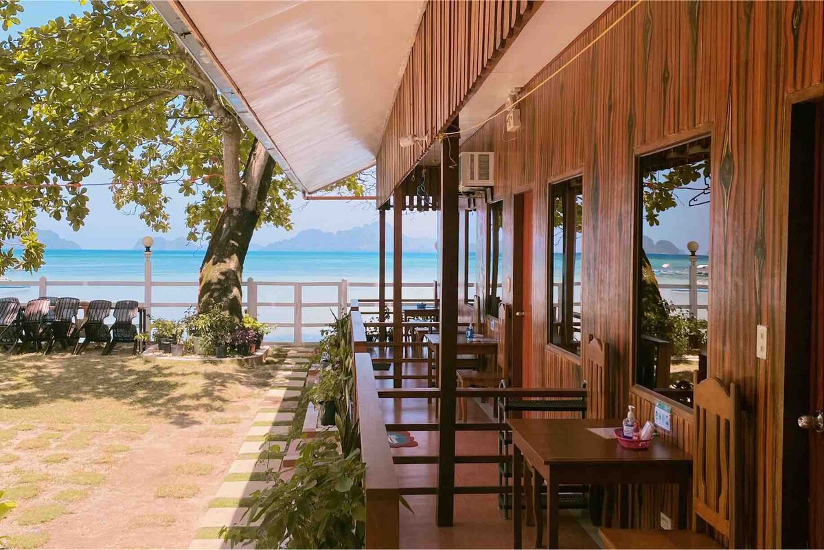 A shaded patio area features wooden tables and chairs, offering views of the beach and sea. Large windows adjacent to the seating area reflect the coastal scenery, while lush greenery surrounds the space, creating a welcoming environment near the water.
