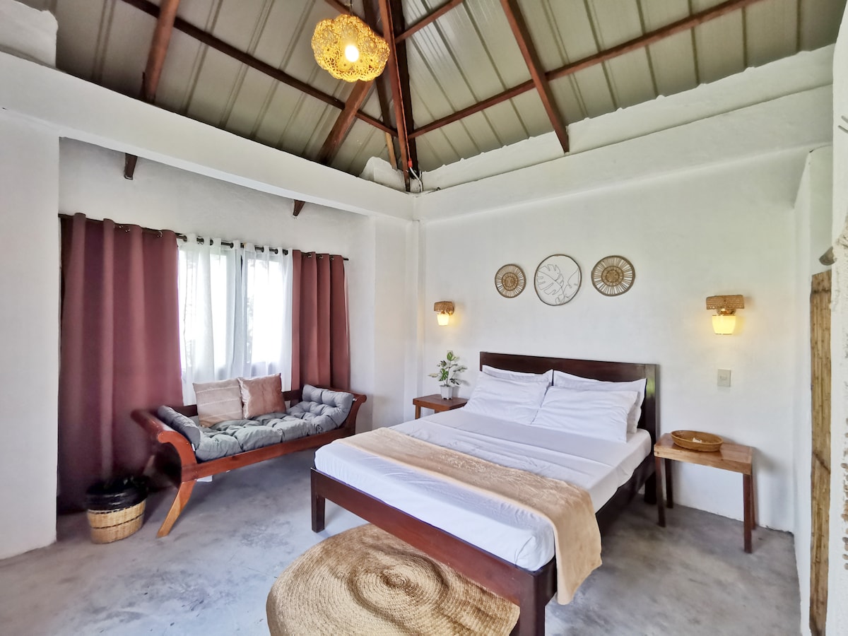 The room features a spacious queen bed with a light-colored bedspread and decorative pillows. A day bed sits against the wall, complemented by natural light from the windows covered by sheer curtains. Wall art and wooden accents enhance the minimalist decor.
