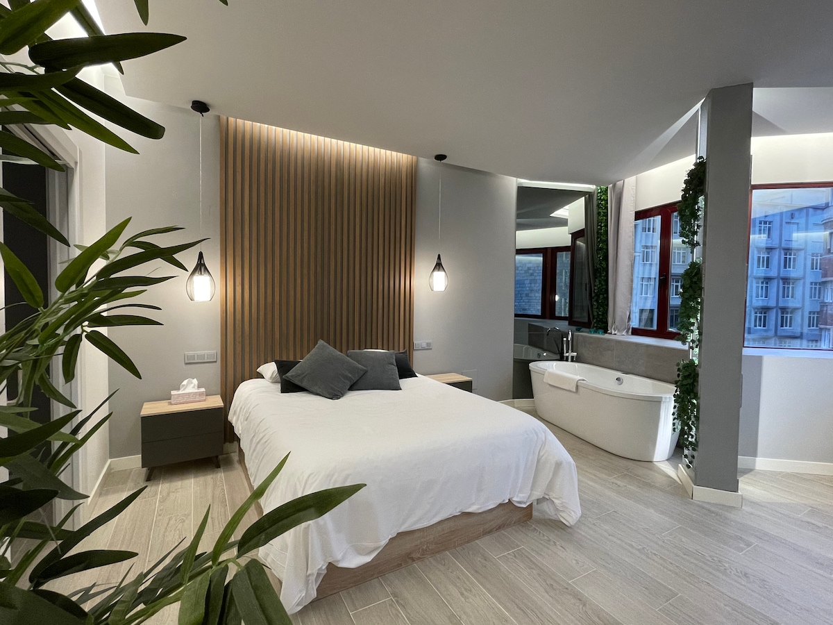 The main room features a spacious bed, framed by a wooden accent wall. A modern freestanding bathtub is visible in the corner, complemented by greenery. Soft lighting is provided by pendant fixtures, enhancing the room's aesthetic without direct sunlight intrusion.