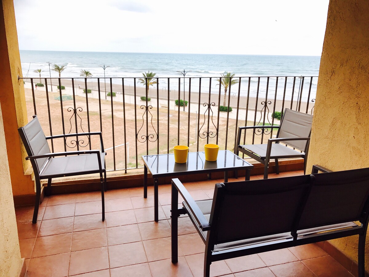 A private balcony offers unobstructed views of the beach and the sea. Two chairs and a loveseat are arranged around a small table. Potted plants are situated on the edge, complementing the serene coastal atmosphere.