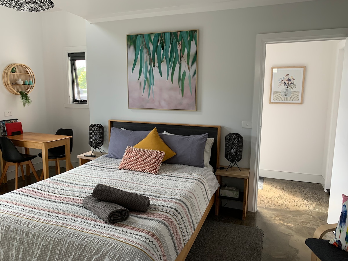 A comfortable sleeping area features a queen bed with patterned bedding and decorative cushions. A dining table with chairs is positioned nearby, while a small ensuite is accessible through a sliding door. Natural light enhances the space, highlighting the polished concrete floor.