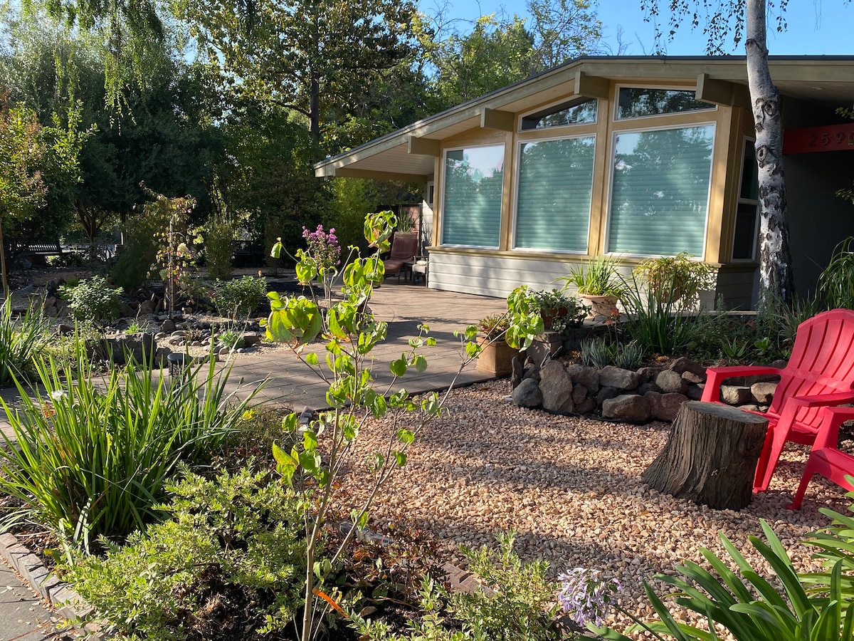 A welcoming front yard showcases a well-maintained garden with native plants and decorative stones. Colorful foliage and flowering plants enhance the natural beauty. A cozy seating area is visible, featuring two red chairs, surrounded by greenery and a pathway leading to the entrance of the home.