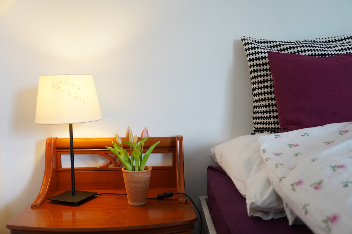 A bedside table is placed next to a spacious bed, featuring a small lamp that casts a warm light. A potted plant with green leaves and pink flowers adds a touch of color to the surface, complementing the patterned pillow and bedding.